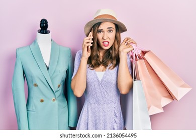 Young Hispanic Girl Holding Shopping Bags And Talking On The Smartphone Clueless And Confused Expression. Doubt Concept. 