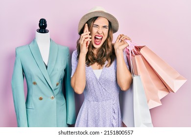 Young Hispanic Girl Holding Shopping Bags And Talking On The Smartphone Angry And Mad Screaming Frustrated And Furious, Shouting With Anger. Rage And Aggressive Concept. 