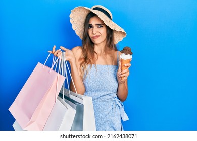 Young Hispanic Girl Holding Shopping Bags And Ice Cream Clueless And Confused Expression. Doubt Concept. 