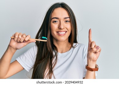 Joven hispana sosteniendo cepillo de dientes con pasta dental sonriendo con una idea o pregunta señalando el dedo con cara feliz, número uno 