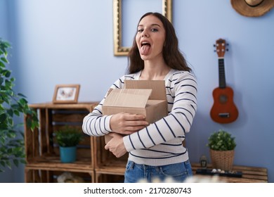 Young Hispanic Girl Opening Cardboard Box Sticking Tongue Out Happy With Funny Expression. 