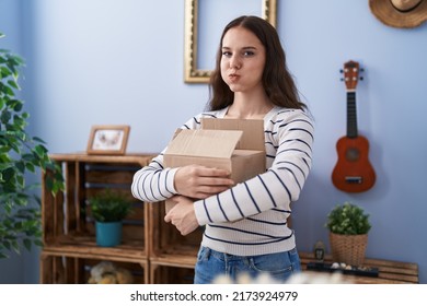 Young Hispanic Girl Opening Cardboard Box Puffing Cheeks With Funny Face. Mouth Inflated With Air, Catching Air. 