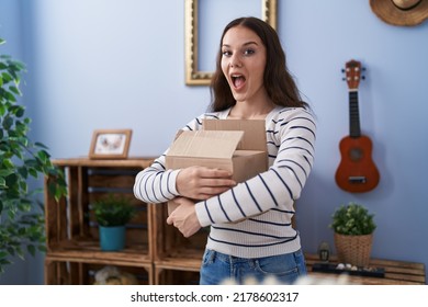 Young Hispanic Girl Opening Cardboard Box Celebrating Crazy And Amazed For Success With Open Eyes Screaming Excited. 