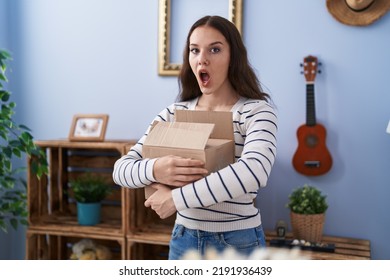 Young Hispanic Girl Opening Cardboard Box Afraid And Shocked With Surprise And Amazed Expression, Fear And Excited Face. 