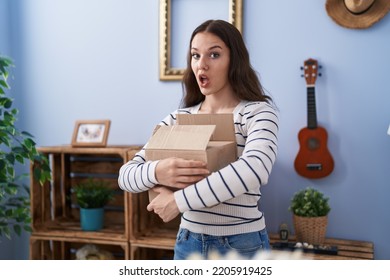 Young Hispanic Girl Opening Cardboard Box In Shock Face, Looking Skeptical And Sarcastic, Surprised With Open Mouth 