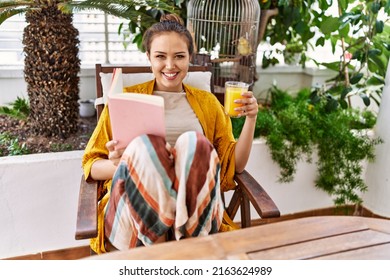 Young Hispanic Girl Reading Book And Drinking Orange Juice At The Terrace.