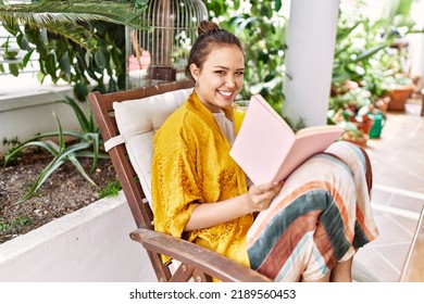 Young Hispanic Girl Reading Book And Drinking Orange Juice At The Terrace.