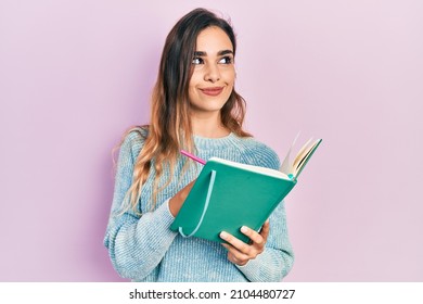 Young hispanic girl reading and writing book smiling looking to the side and staring away thinking. 