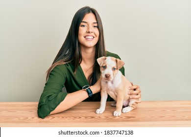 Young Hispanic Girl Smiling Happy And Playing With Dog Sitting On The Table Over Isolated White Background.