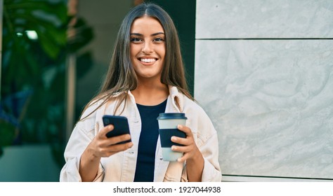 Young hispanic girl smiling happy using smartphone and drinking take away coffee at the city.