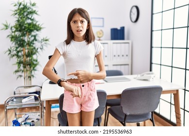 Young hispanic girl standing at pediatrician clinic in hurry pointing to watch time, impatience, upset and angry for deadline delay 