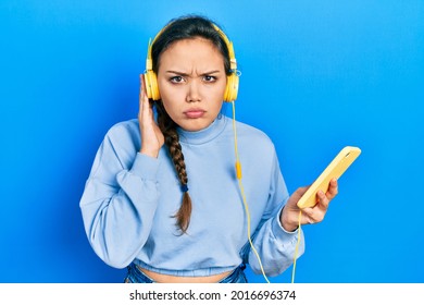 Young Hispanic Girl Using Smartphone And Headphones Skeptic And Nervous, Frowning Upset Because Of Problem. Negative Person. 