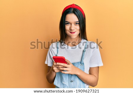 Young hispanic girl using smartphone puffing cheeks with funny face. mouth inflated with air, catching air. 