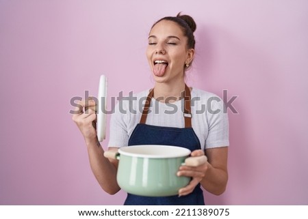 Young hispanic girl wearing apron holding cooking pot sticking tongue out happy with funny expression. 