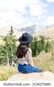 Young Hispanic girl wearing a black fedora hat siting on a rock enjoying a beautiful mountain scenery 