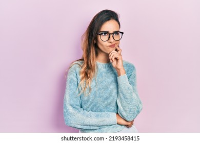 Young Hispanic Girl Wearing Casual Clothes And Glasses Serious Face Thinking About Question With Hand On Chin, Thoughtful About Confusing Idea 