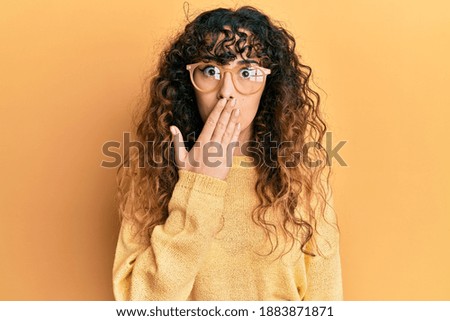 Young hispanic girl wearing casual clothes and glasses covering mouth with hand, shocked and afraid for mistake. surprised expression 