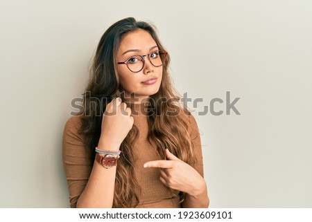 Young hispanic girl wearing casual clothes and glasses in hurry pointing to watch time, impatience, looking at the camera with relaxed expression 