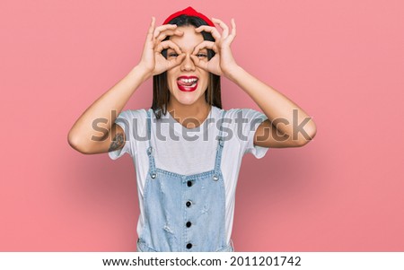 Young hispanic girl wearing casual clothes doing ok gesture like binoculars sticking tongue out, eyes looking through fingers. crazy expression. 
