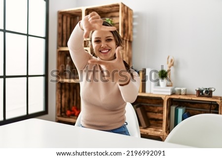 Young hispanic girl wearing casual clothes sitting on the table at home smiling making frame with hands and fingers with happy face. creativity and photography concept. 