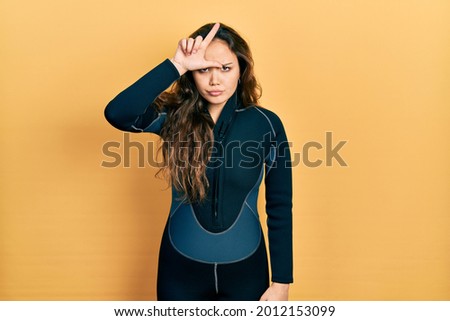 Young hispanic girl wearing diver neoprene uniform making fun of people with fingers on forehead doing loser gesture mocking and insulting. 