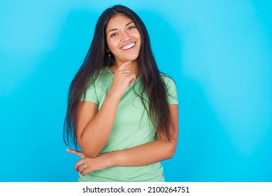 Young hispanic girl wearing green T-shirt over blue background laughs happily keeps hand on chin expresses positive emotions smiles broadly has carefree expression