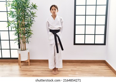 Young hispanic girl wearing karate kimono and black belt making fish face with lips, crazy and comical gesture. funny expression. 