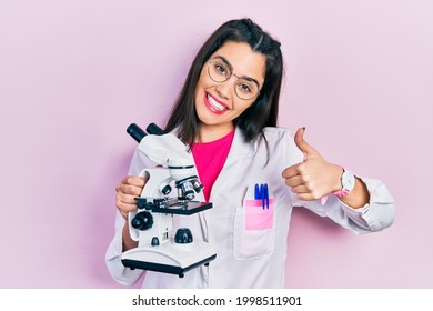 Young Hispanic Girl Wearing Scientist Uniform Holding Microscope Smiling Happy And Positive, Thumb Up Doing Excellent And Approval Sign 