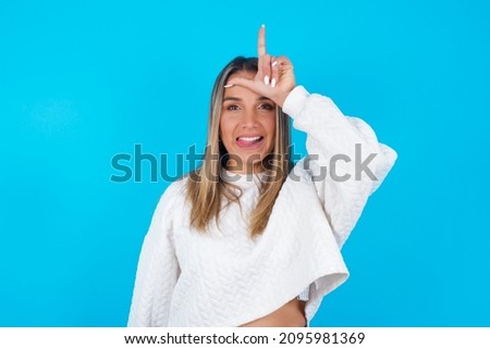 Young hispanic girl wearing white knitted sweater over blue background gestures with finger on forehead makes loser gesture makes fun of people shows tongue