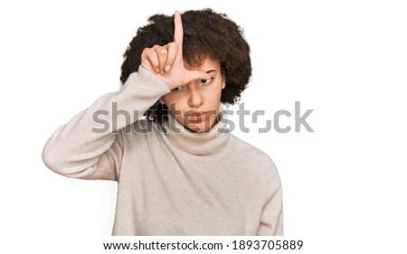 Young hispanic girl wearing wool winter sweater making fun of people with fingers on forehead doing loser gesture mocking and insulting. 