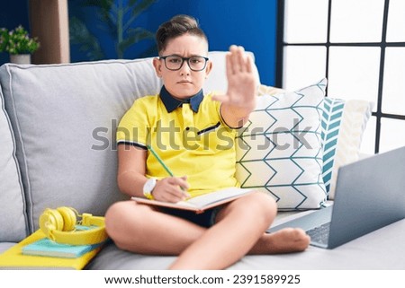 Young hispanic kid doing homework sitting on the sofa doing stop sing with palm of the hand. warning expression with negative and serious gesture on the face. 