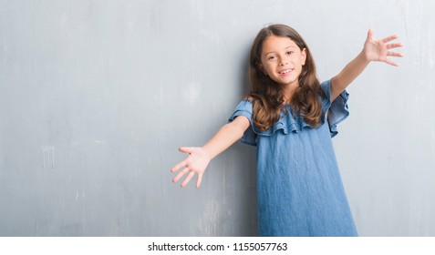 Young Hispanic Kid Over Grunge Grey Wall Looking At The Camera Smiling With Open Arms For Hug. Cheerful Expression Embracing Happiness.