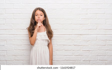 Young Hispanic Kid Over White Brick Wall Asking To Be Quiet With Finger On Lips. Silence And Secret Concept.