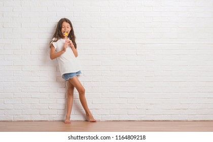 Young hispanic kid over white brick wall eating lollipop candy at home with a confident expression on smart face thinking serious