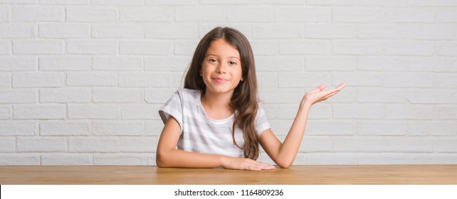 Young Hispanic Kid Sitting On The Table At Home Smiling Cheerful Presenting And Pointing With Palm Of Hand Looking At The Camera.