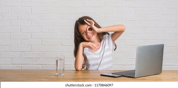 Young Hispanic Kid Sitting On The Table Using Computer Laptop With Happy Face Smiling Doing Ok Sign With Hand On Eye Looking Through Fingers