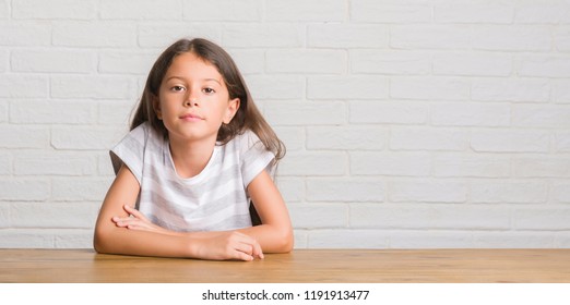 Young Hispanic Kid Sitting On The Table At Home With Serious Expression On Face. Simple And Natural Looking At The Camera.