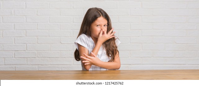 Young Hispanic Kid Sitting On The Table At Home Smelling Something Stinky And Disgusting, Intolerable Smell, Holding Breath With Fingers On Nose. Bad Smells Concept.