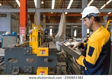Young hispanic labor dressed in polyester jacket uniform, hardhat and protective glove check the orderliness of the machine. Working atmosphere in metal sheet factory with large industrial machines.