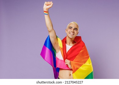 Young hispanic latin gay man 20s with make up wrapped in rainbow striped flag raise up clench fist look overhead isolated on plain pastel purple background. People lifestyle fashion lgbtq concept.