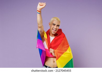 Young hispanic latin gay man 20s with make up wrapped in rainbow striped flag raise up clench fist isolated on plain pastel purple background studio portrait. People lifestyle fashion lgbtq concept.
