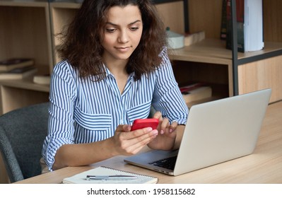 Young Hispanic Latin Student Holding Smartphone Studying On Cell And Laptop, Using Mobile Virtual Learning Classes Tech Apps. Teenage Female Indian Girl Watching Online Training At Creative Office.