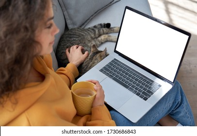 Young hispanic latin teen girl student relax sit on sofa with cat holding laptop looking at mock up white computer screen online learning on pc, elearning, watching movie. Over shoulder closeup view