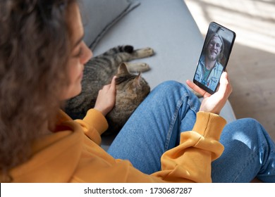Young hispanic latin teen girl relax sit on sofa with cat at home holding phone video calling distance friend dating online on mobile screen using smartphone videochat app. Over shoulder closeup view