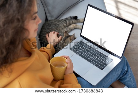 Young hispanic latin teen girl student relax sit on sofa with cat holding laptop looking at mock up white computer screen online learning on pc, elearning, watching movie. Over shoulder closeup view