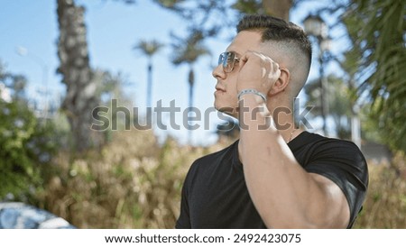 A young hispanic man adjusts his sunglasses in a sunny park with palm trees.