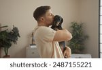 A young hispanic man affectionately holds and kisses his dachshund dog in a cozy living room, surrounded by plants and soft lighting, capturing a tender moment of pet love and companionship.