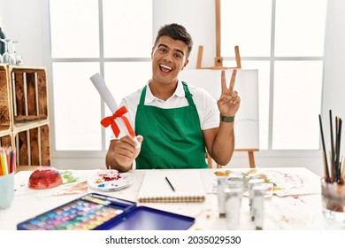 Young hispanic man at art studio holding degree smiling with happy face winking at the camera doing victory sign with fingers. number two. 