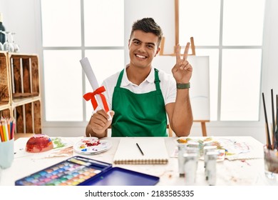 Young hispanic man at art studio holding degree smiling looking to the camera showing fingers doing victory sign. number two. 