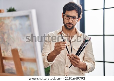 Young hispanic man artist holding paintbrushes with serious expression at art studio